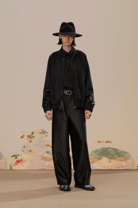 A model wearing a black bomber jacket with wide black trousers and a wide-brim hat
