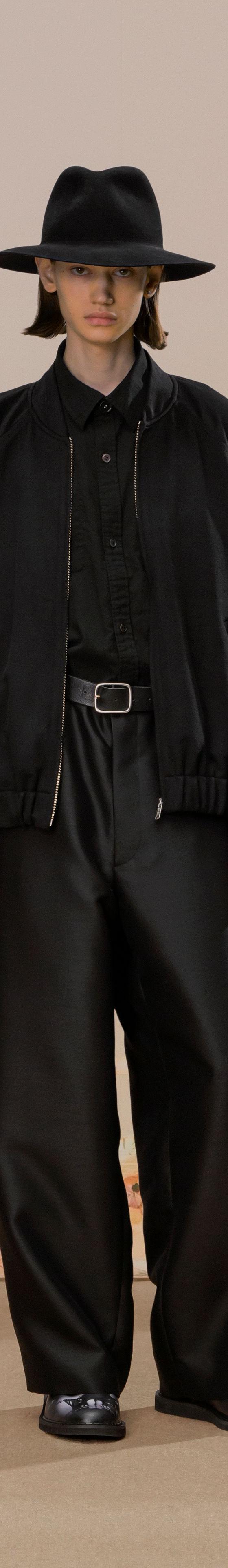 A model wearing a black bomber jacket with wide black trousers and a wide-brim hat