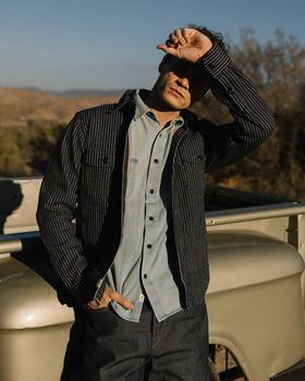 A man wearing a black striped Hiroshi Kato work jacket over a light blue denim shirt and indigo jeans, leaning against a vintage truck