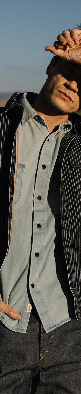 A man wearing a black striped Hiroshi Kato work jacket over a light blue denim shirt and indigo jeans, leaning against a vintage truck