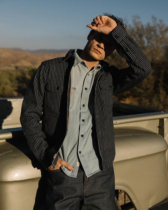 A man wearing a black striped Hiroshi Kato work jacket over a light blue denim shirt and indigo jeans, leaning against a vintage truck