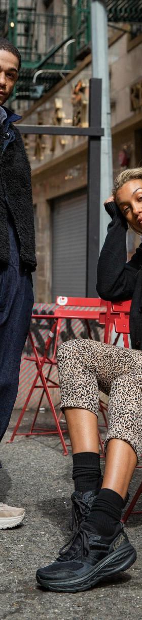 Two people wearing HOKA sneakers with layered streetwear on a city sidewalk