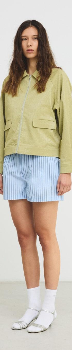 A woman wearing a pistachio green Hosbjerg jacket with blue striped shorts and white sandals