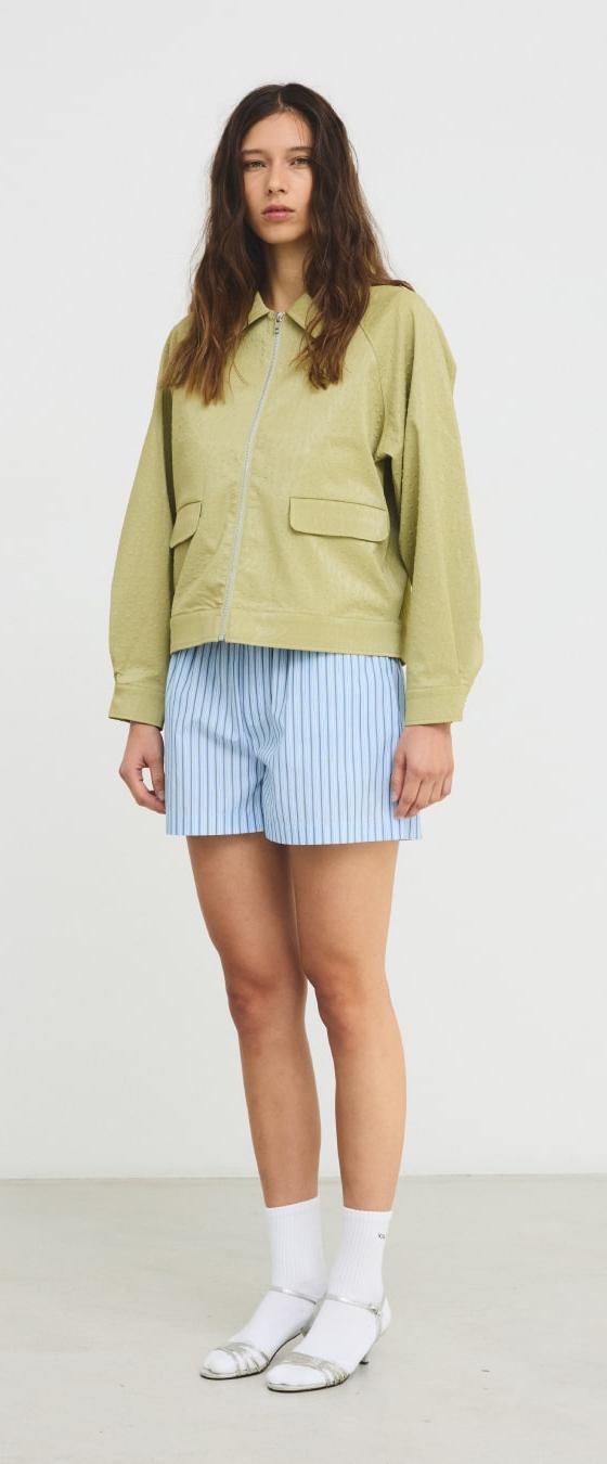 A woman wearing a pistachio green Hosbjerg jacket with blue striped shorts and white sandals