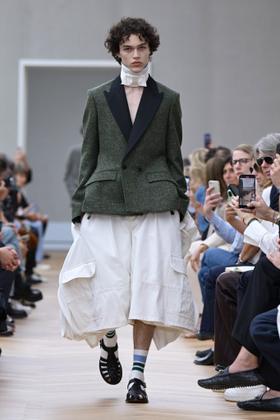 A man wearing a green tweed tuxedo-lapel jacket with wide white culottes