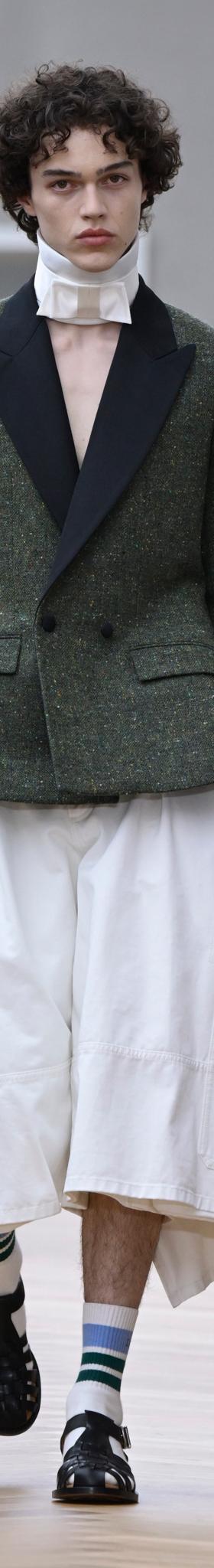 A man wearing a green tweed tuxedo-lapel jacket with wide white culottes