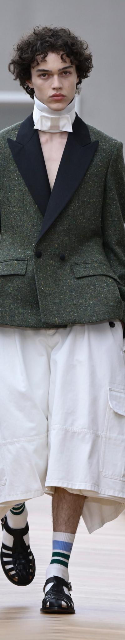 A man wearing a green tweed tuxedo-lapel jacket with wide white culottes