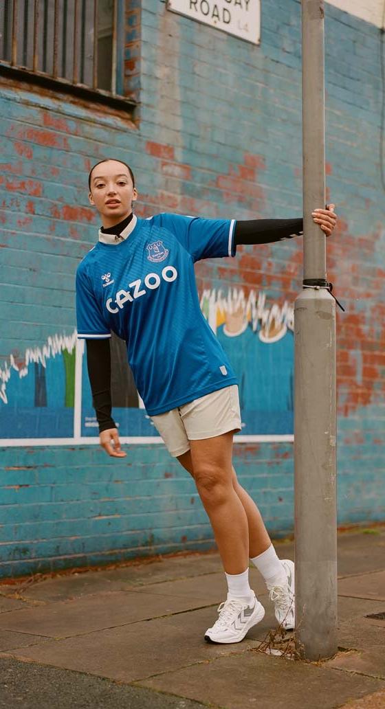 A woman wearing a blue hummel Everton football shirt with white shorts and sneakers on a city street