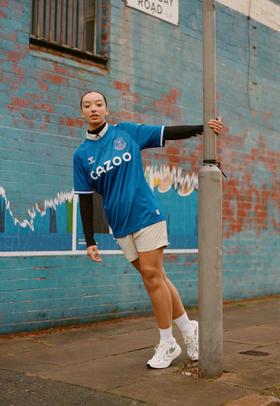 A woman wearing a blue hummel Everton football shirt with white shorts and sneakers on a city street