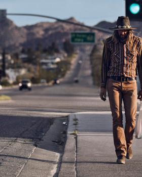 A man in a striped western shirt, felt hat and tan jeans walking along a desert highway