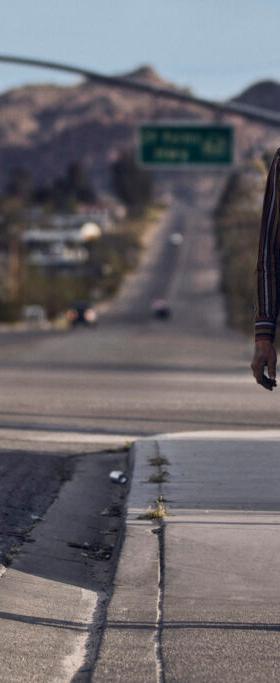 A man in a striped western shirt, felt hat and tan jeans walking along a desert highway