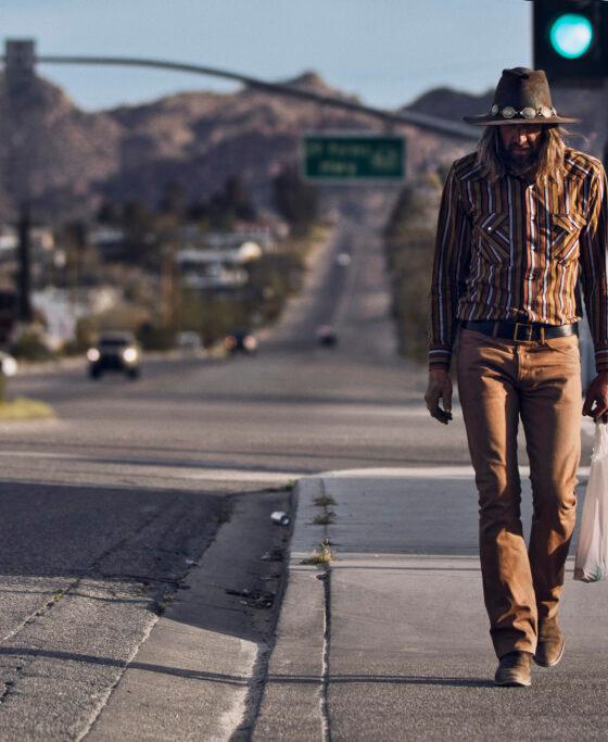 A man in a striped western shirt, felt hat and tan jeans walking along a desert highway