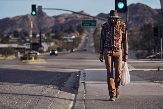 A man in a striped western shirt, felt hat and tan jeans walking along a desert highway