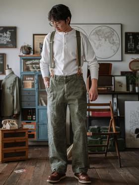 A man wearing an INDIGOSEA white henley with olive suspenders and vintage military-style trousers