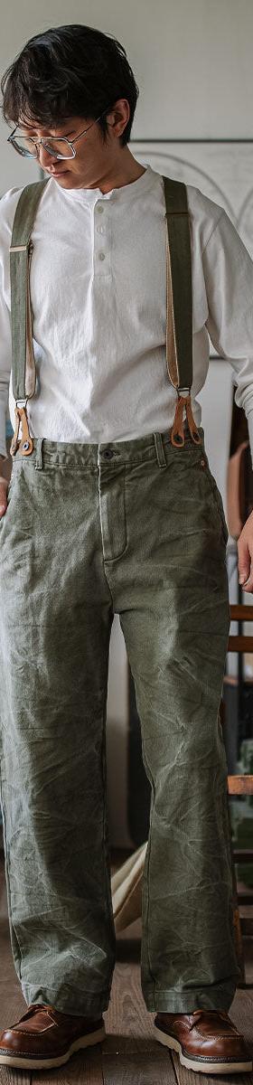 A man wearing an INDIGOSEA white henley with olive suspenders and vintage military-style trousers