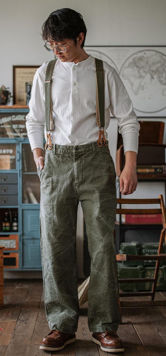 A man wearing an INDIGOSEA white henley with olive suspenders and vintage military-style trousers
