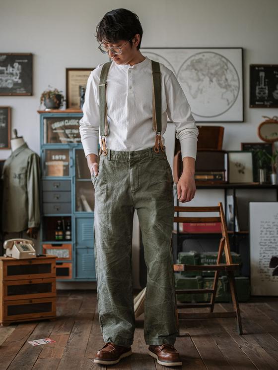 A man wearing an INDIGOSEA white henley with olive suspenders and vintage military-style trousers