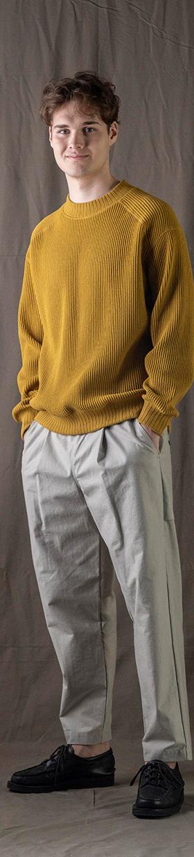 A man wearing a mustard yellow Jackman knit sweater with light grey pleated trousers
