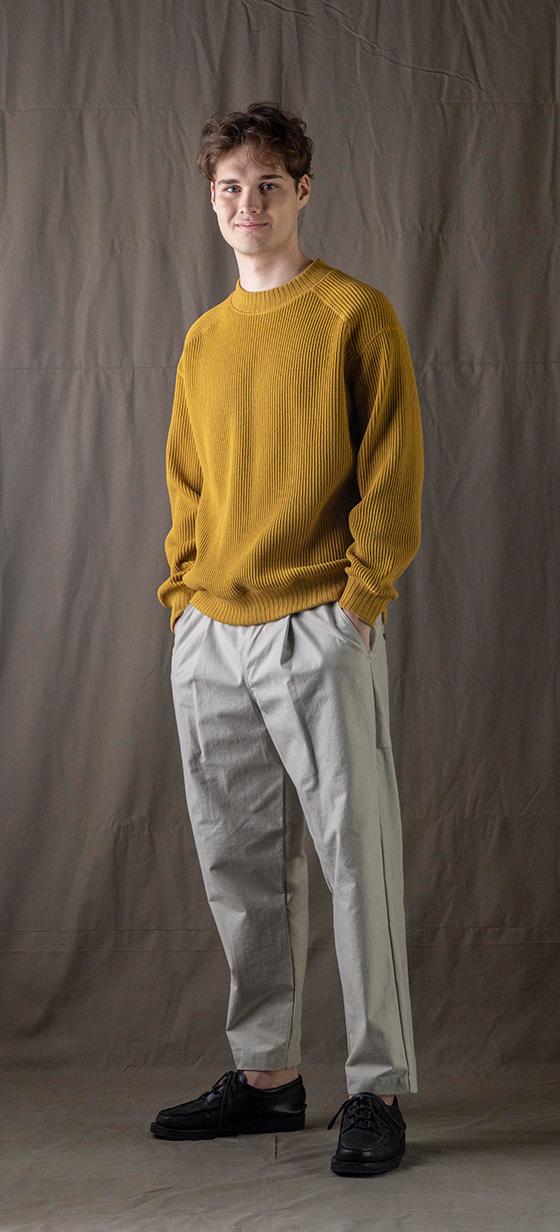 A man wearing a mustard yellow Jackman knit sweater with light grey pleated trousers