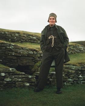 A man wearing a dark green John Alexander Skelton coat and trousers with a rope-tied waistcoat, standing on a grassy hillside