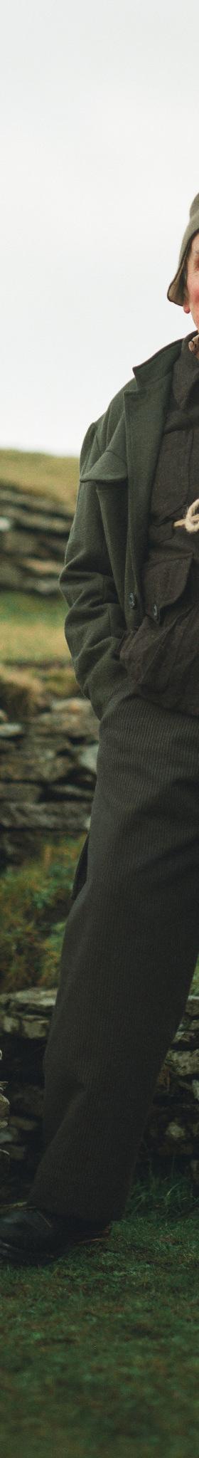 A man wearing a dark green John Alexander Skelton coat and trousers with a rope-tied waistcoat, standing on a grassy hillside