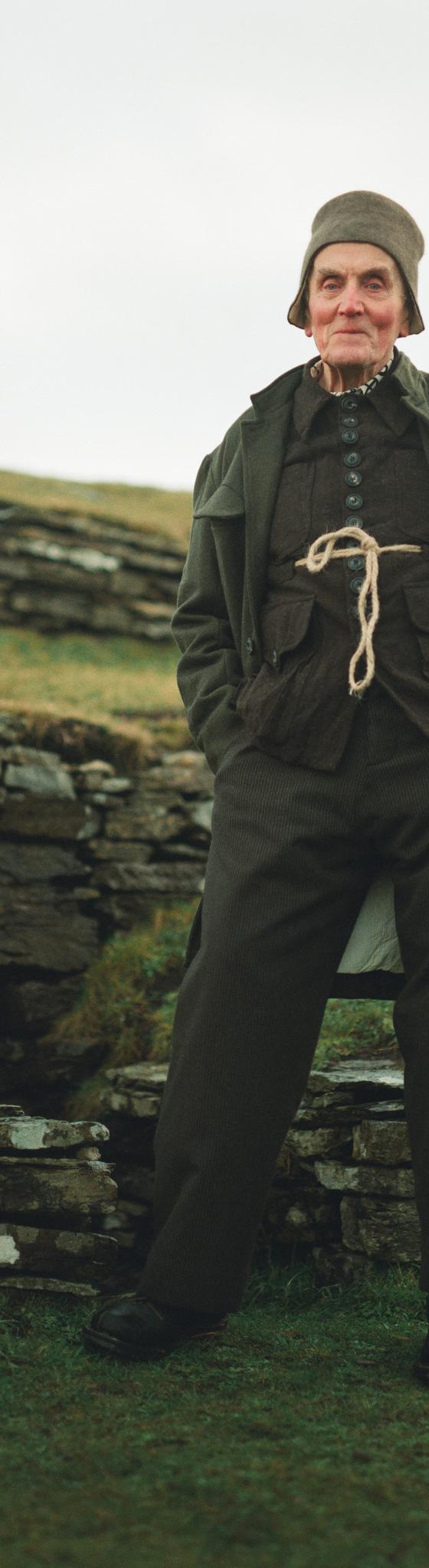 A man wearing a dark green John Alexander Skelton coat and trousers with a rope-tied waistcoat, standing on a grassy hillside
