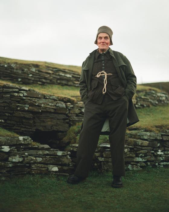 A man wearing a dark green John Alexander Skelton coat and trousers with a rope-tied waistcoat, standing on a grassy hillside