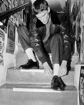 A man in a leather jacket and jeans sitting on stairs lacing black John Lobb leather derby shoes