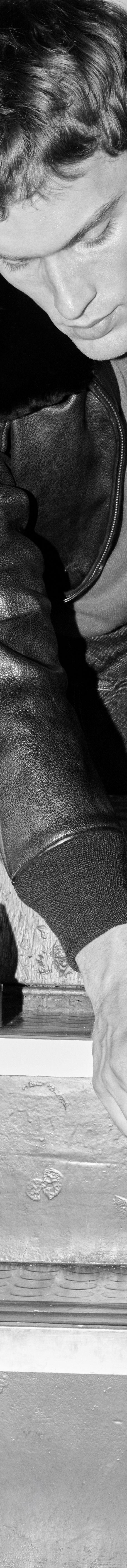 A man in a leather jacket and jeans sitting on stairs lacing black John Lobb leather derby shoes