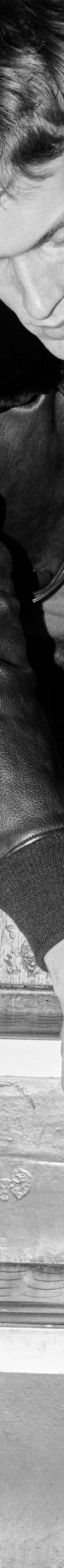 A man in a leather jacket and jeans sitting on stairs lacing black John Lobb leather derby shoes