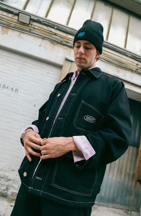 A man wearing a black Kaotiko Society denim chore jacket with contrast stitching and a black beanie