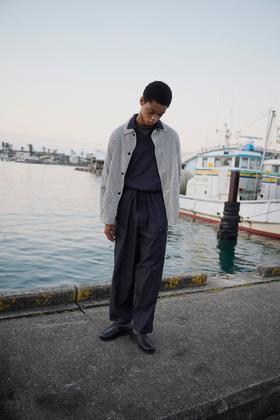 A man wearing a striped Kaptain Sunshine coach jacket with wide navy trousers on a harbour dock