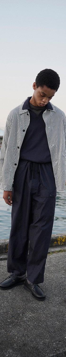 A man wearing a striped Kaptain Sunshine coach jacket with wide navy trousers on a harbour dock