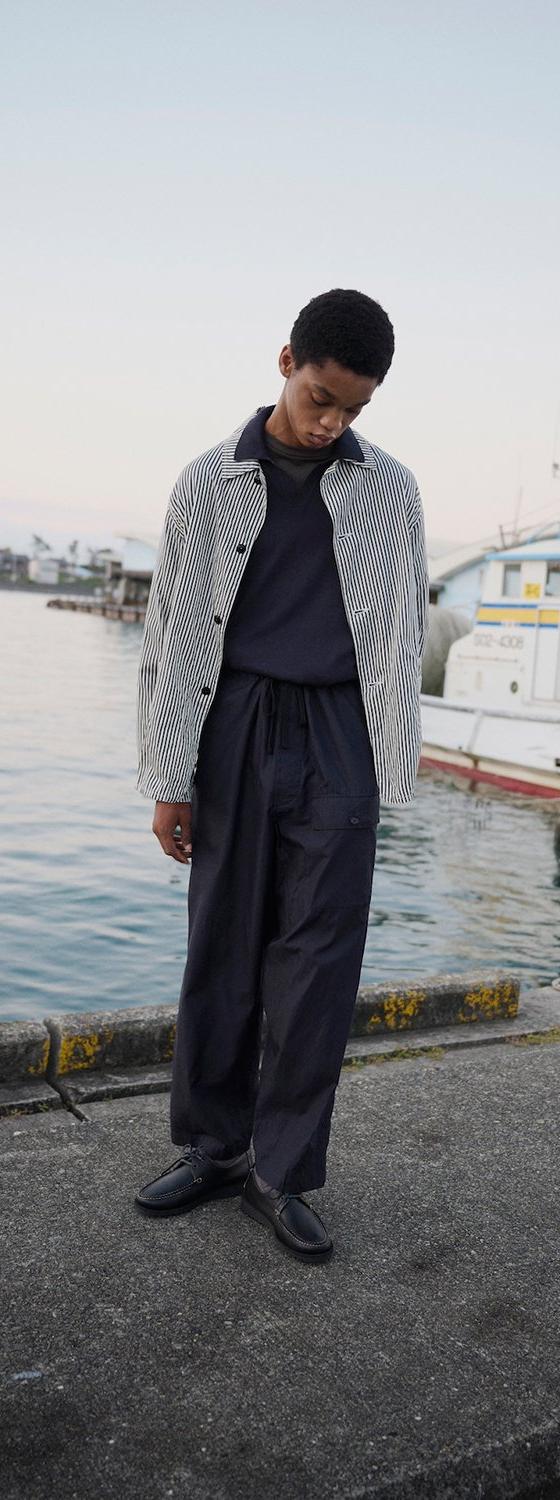 A man wearing a striped Kaptain Sunshine coach jacket with wide navy trousers on a harbour dock