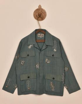 A sage green Kartik Research chore jacket with hand-embroidered motifs, hung on a wooden hanger