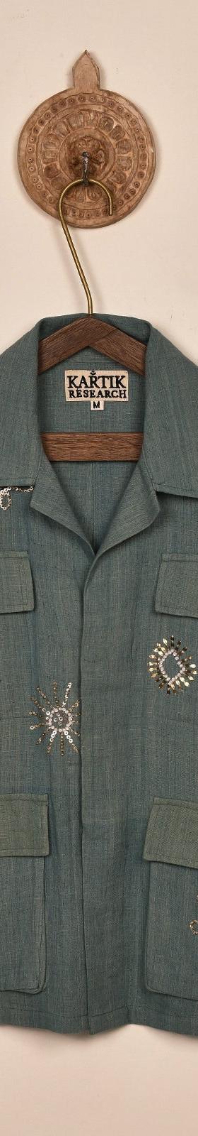 A sage green Kartik Research chore jacket with hand-embroidered motifs, hung on a wooden hanger