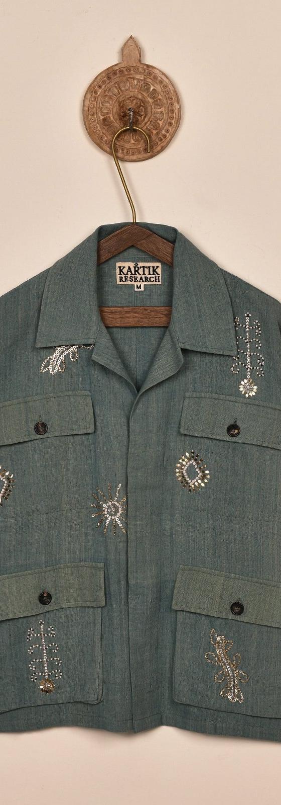 A sage green Kartik Research chore jacket with hand-embroidered motifs, hung on a wooden hanger