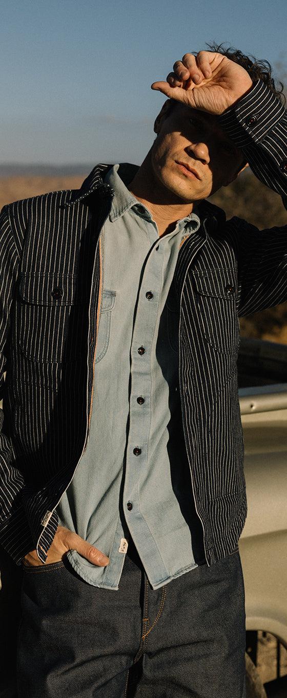 A man wearing a black striped KATO work jacket over a light blue denim shirt and dark selvedge jeans, leaning against a vintage pickup truck