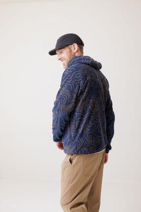 A man wearing a navy topo-print KAVU fleece anorak with a black cap and tan trousers