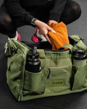 A woman crouching beside an olive green KNKG gym duffel bag on a gym floor