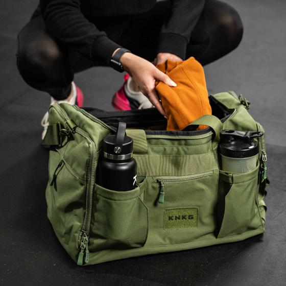 A woman crouching beside an olive green KNKG gym duffel bag on a gym floor