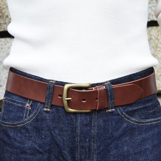 A person wearing a brown leather Kobashi Studio belt with a solid-brass buckle over indigo denim