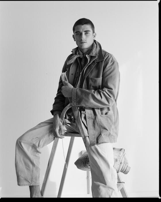 A man wearing a herringbone L.E.J chore jacket with a neckerchief and light trousers, seated on a stepladder