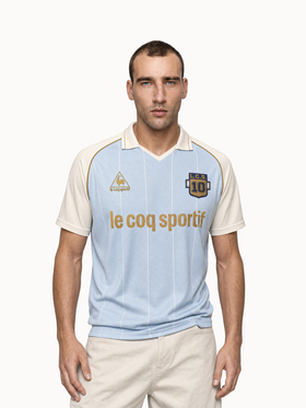A man wearing a light blue Le Coq Sportif retro football jersey with cream sleeves
