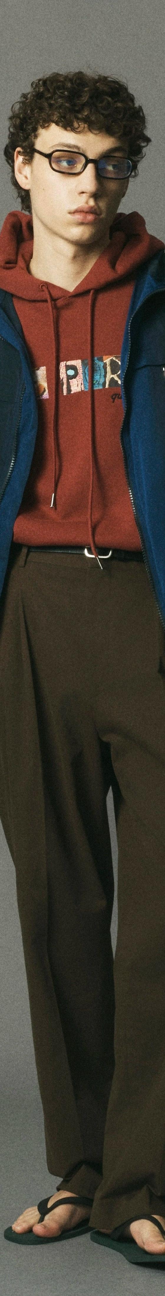 A man wearing a blue le PÈRE hooded jacket over a rust graphic hoodie with brown trousers