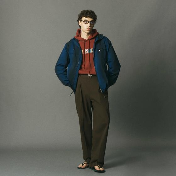 A man wearing a blue le PÈRE hooded jacket over a rust graphic hoodie with brown trousers