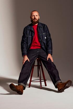 A bearded man wearing a raw indigo Lee 101 denim jacket and selvedge jeans with brown leather boots, seated on a stool