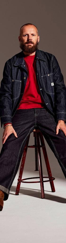 A bearded man wearing a raw indigo Lee 101 denim jacket and selvedge jeans with brown leather boots, seated on a stool