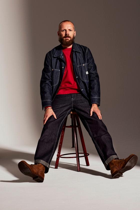 A bearded man wearing a raw indigo Lee 101 denim jacket and selvedge jeans with brown leather boots, seated on a stool