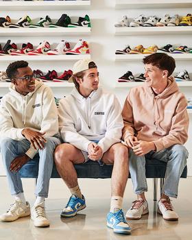 Three men in hoodies and sneakers sitting in a sneaker boutique lined with shelves of Nike and Air Jordan shoes